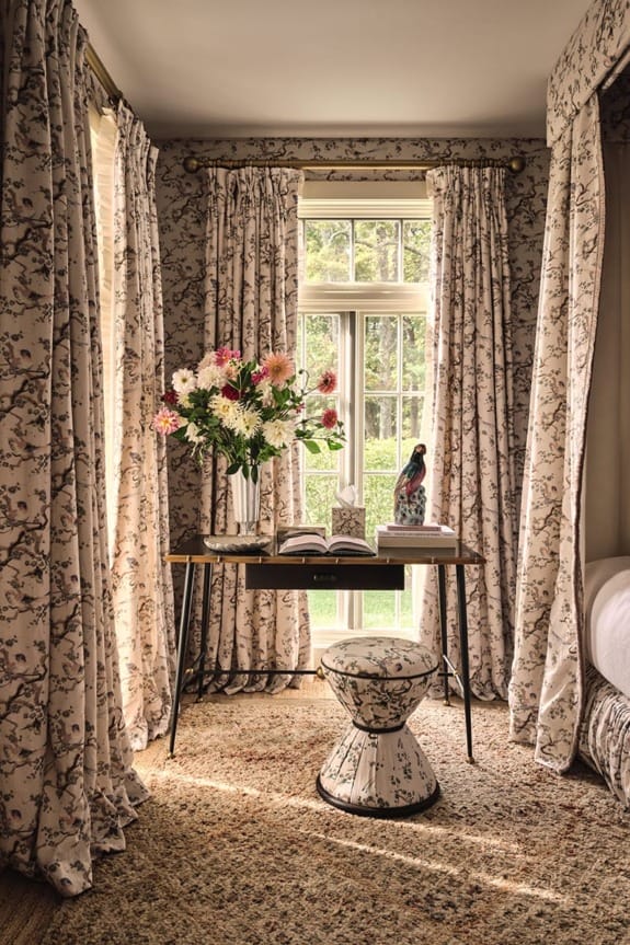 Cozy room with floral curtains, a small desk with a vase of flowers, and a stool, sunlit window in the background.
