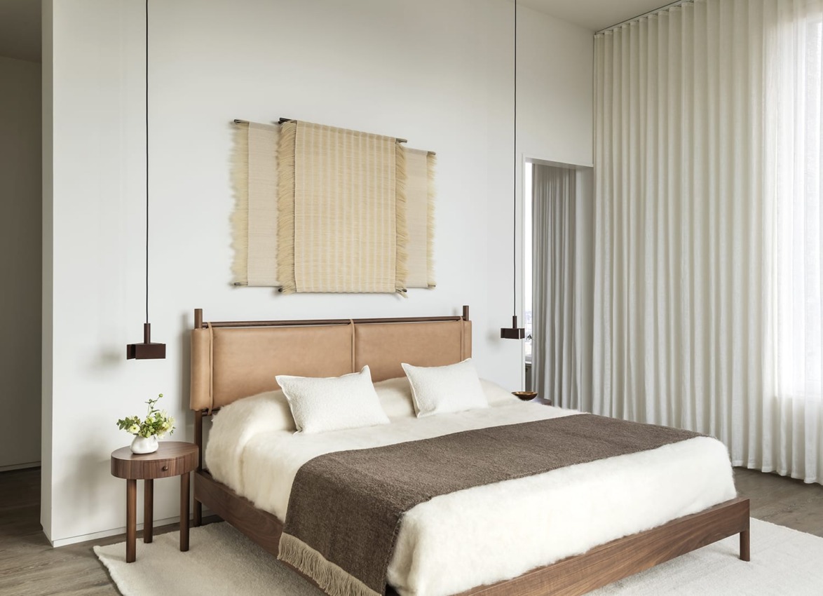 Modern bedroom with a wooden bed, neutral bedding, side table with plant, wall art, and large white curtains.