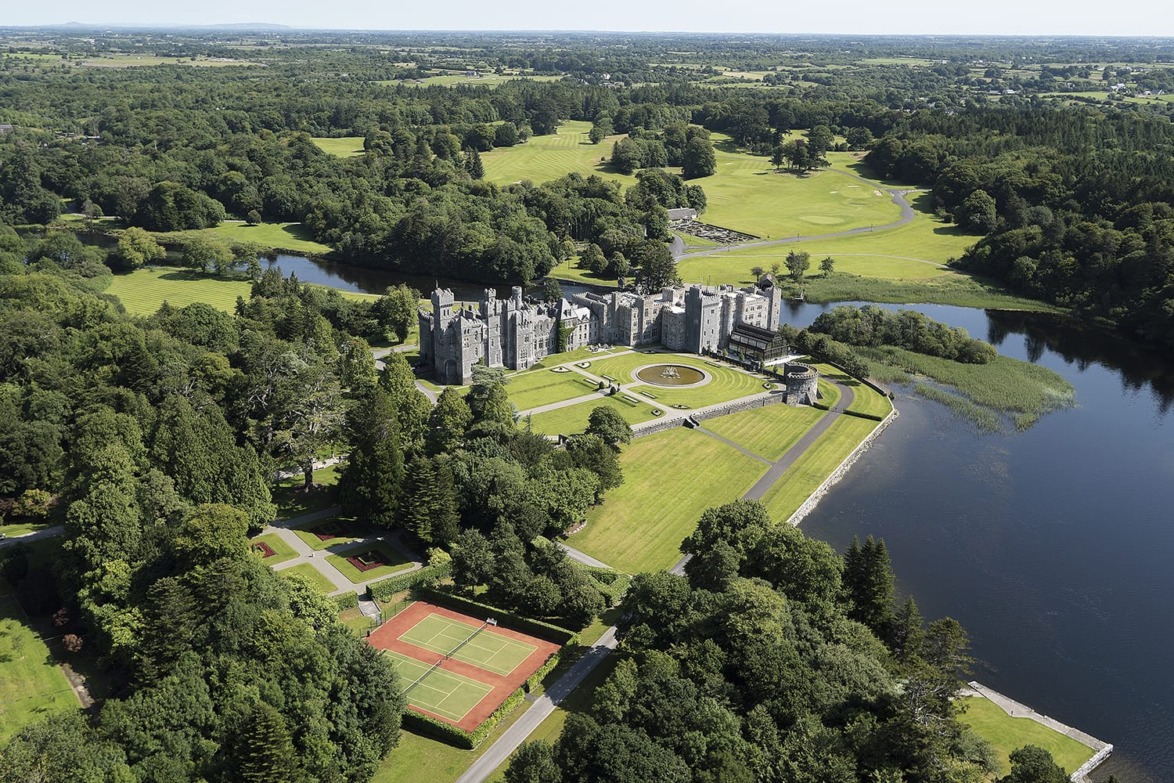 Aerial view of a historic castle surrounded by lush greenery, gardens, and a river with tennis courts nearby.