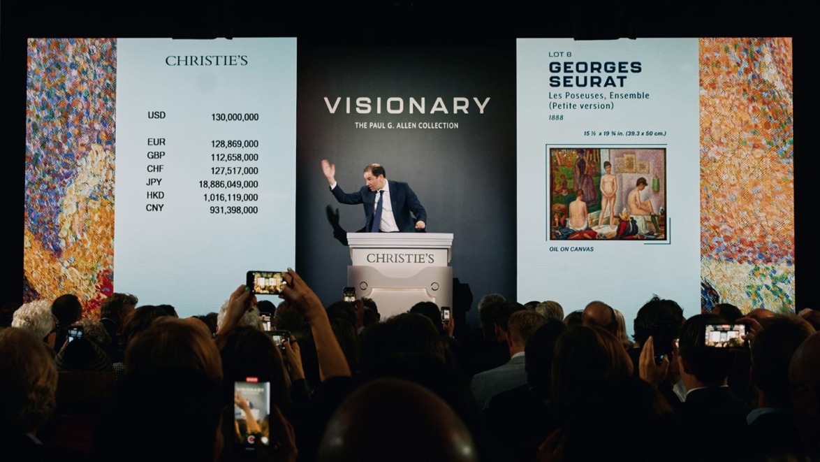 Auctioneer at Christie's sale with art piece displayed and currency bids shown on screen, audience capturing the moment.