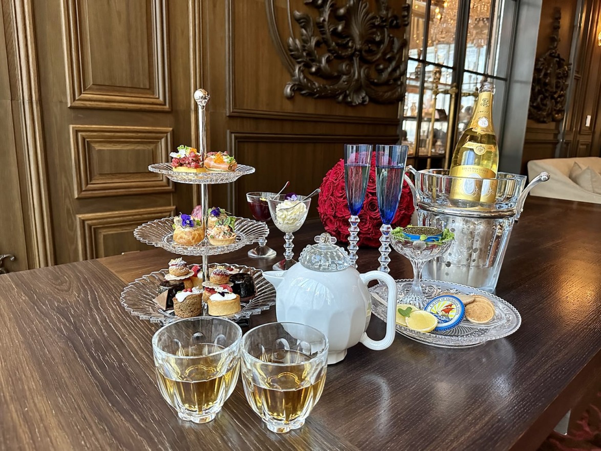 Elegant afternoon tea setup with tiered tray of pastries, teapot, glasses of tea, wine bottle, and ice bucket on wooden table.