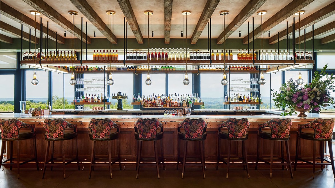 Stylish bar interior with floral-patterned chairs, hanging lights, and a wide selection of colorful bottles on shelves.