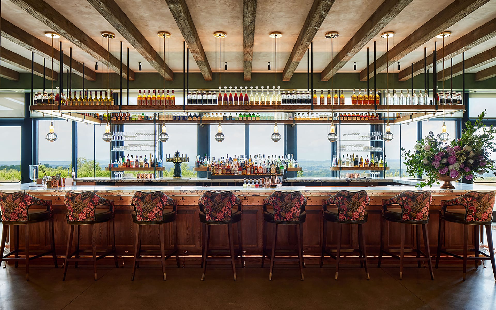 Stylish bar interior with floral-patterned chairs, hanging lights, and a wide selection of colorful bottles on shelves.