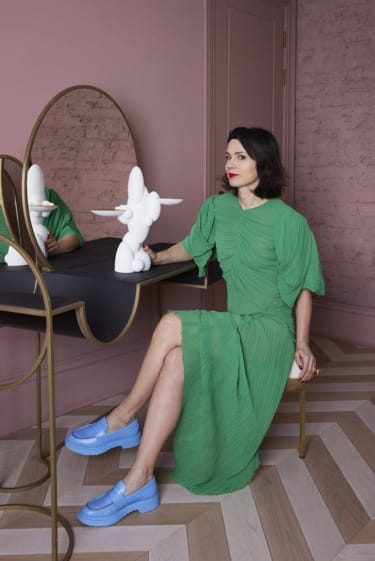Woman in green dress sits by a vanity with abstract sculpture, wearing blue shoes, against a pink wall background.