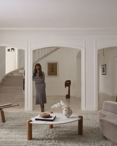 Modern living room with a woman standing near staircase, featuring a gray sofa, white coffee table, and minimalist decor.