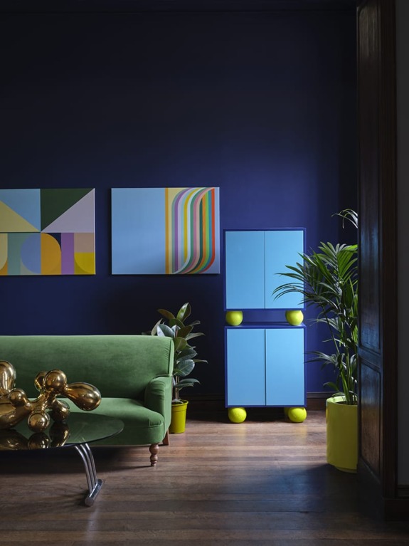 Modern living room with green sofa, abstract art, blue cabinets, and plants in yellow pots against a dark blue wall.