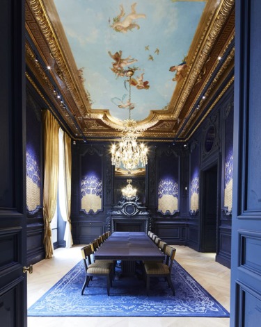 Elegant dining room with ornate ceiling mural, chandeliers, dark walls, golden curtains, and a long table with chairs.