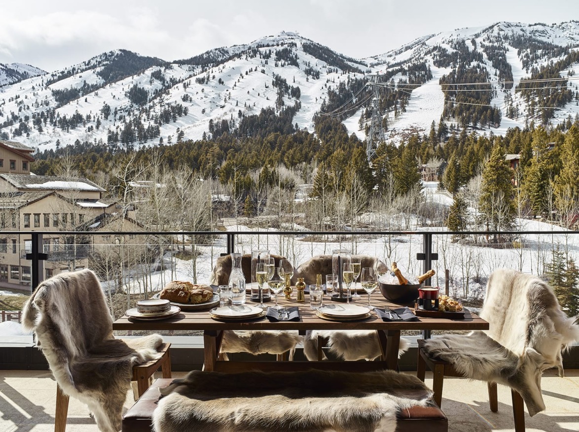 Outdoor dining setup with table, chairs, and food, overlooking snowy mountains and trees.