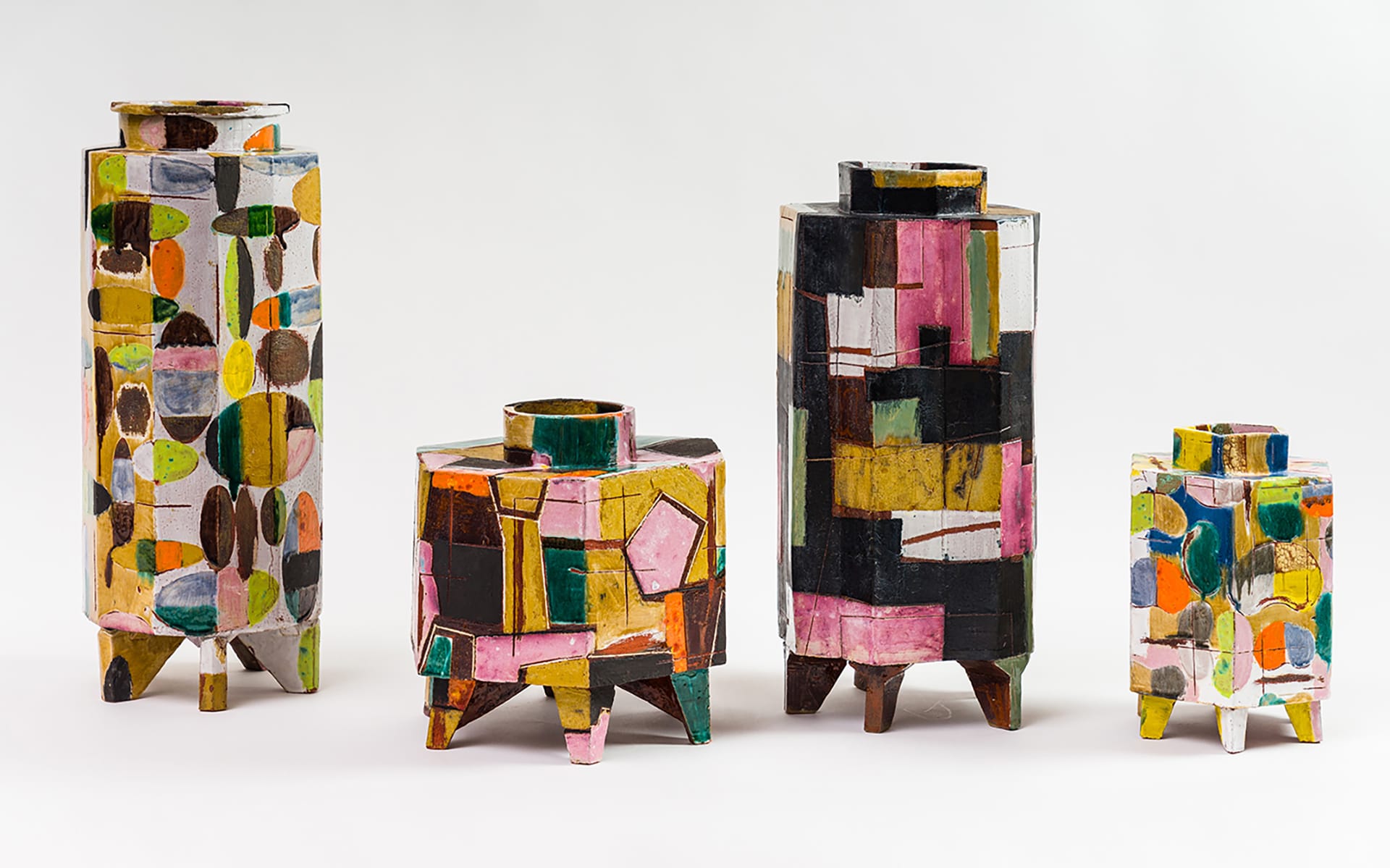 Four colorful, geometric vases with abstract designs and wooden legs arranged in a row against a plain background.