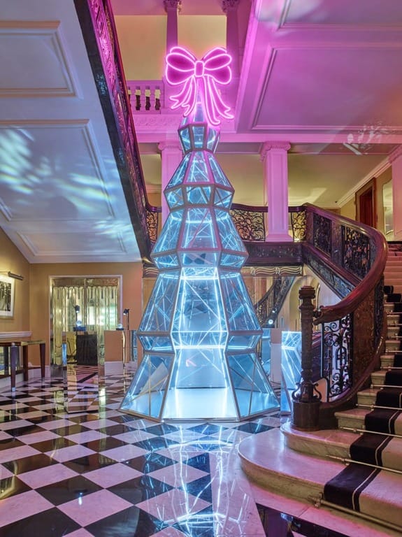 Modern artistic Christmas tree with neon lights displayed in an elegant interior with a checkered floor and grand staircase.