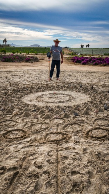 Man standing on a sandy field creating geometric patterns with a stick, mountains and flowers in the background.