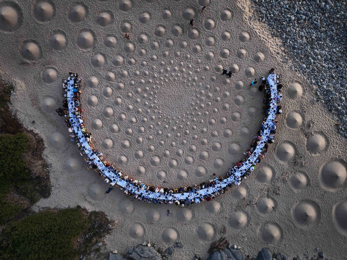 Aerial view of long dinner table set on sandy beach, surrounded by circular sand formations and green foliage.