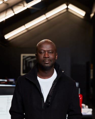 A person in a black jacket stands in a modern office setting with ceiling lights in the background.