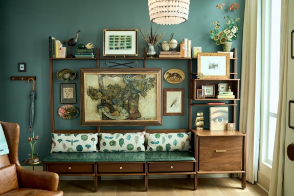 Cozy living room with teal walls, art-filled shelves, wooden furniture, floral cushions, and a chandelier.