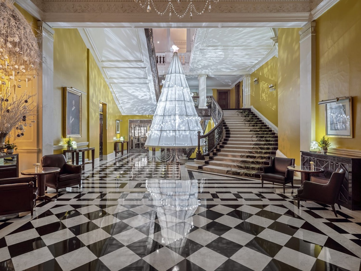 Elegant lobby with a checkered floor, grand staircase, and modern chandelier centerpiece surrounded by seating areas.