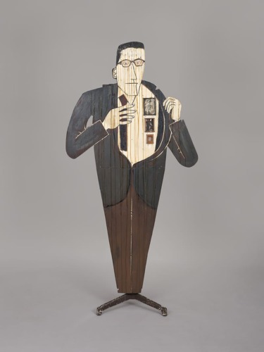 Wooden sculpture of a person in a suit with visible photographs on the chest area against a plain background.