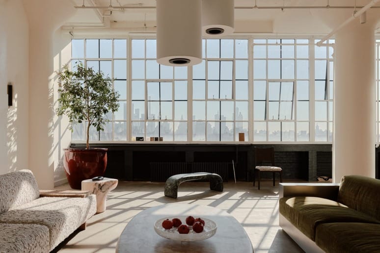 Spacious, sunlit living room with modern furniture, large windows, and city skyline in the background.