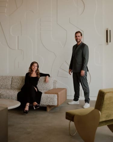 Two people in a modern interior with abstract wall design, one seated on a sofa and the other standing.