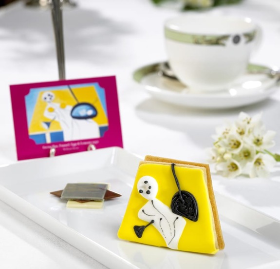 Decorative biscuit with abstract icing art on a white plate, accompanied by a teacup and flowers in a refined setting.