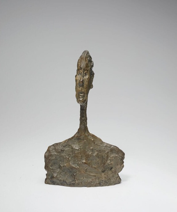 Abstract bronze sculpture of a thin elongated face on a rugged base against a neutral background