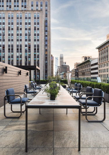Modern rooftop terrace with long table, blue chairs, and cityscape views under a clear sky.