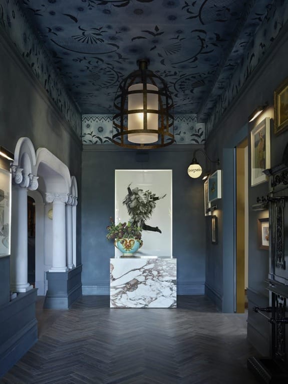 Elegant hallway with dark blue walls, ornate ceiling, large lantern light, marble pedestal, and framed artworks.