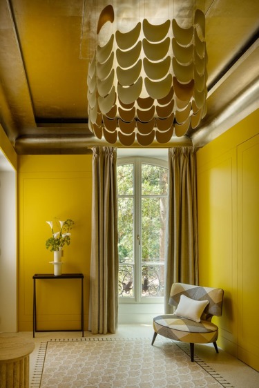 Modern room with bright yellow walls, patterned chair, decorative ceiling light, and a window with curtains and a small table with flowers.