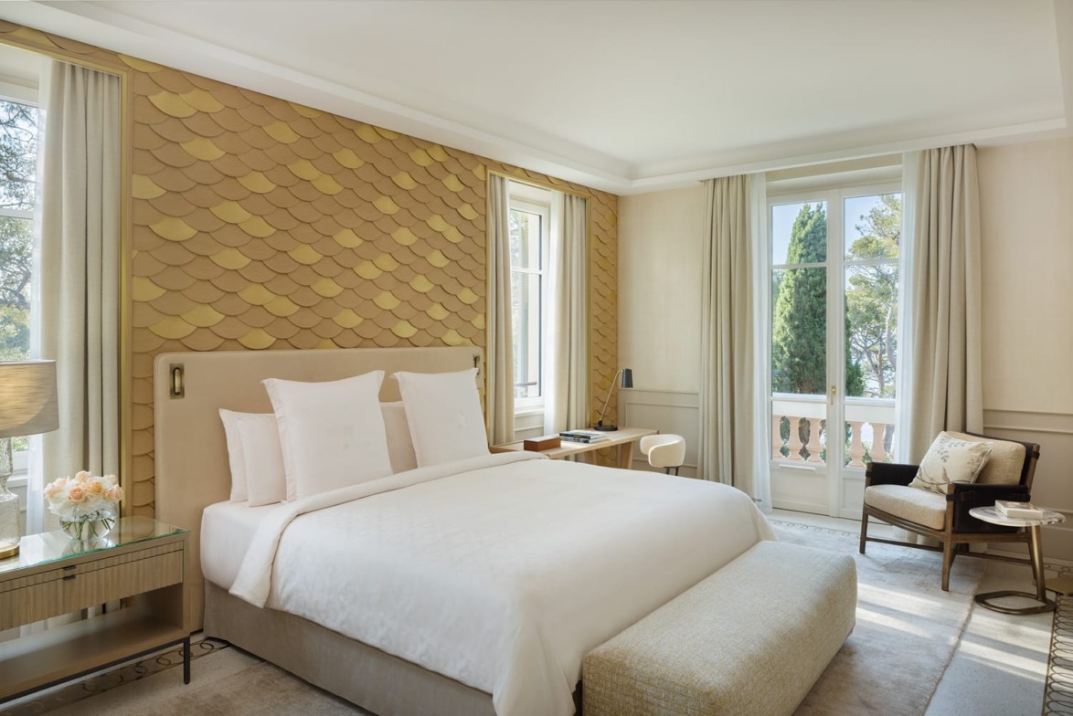 Elegant hotel room with a plush bed, textured wall, large windows, and stylish furniture offering a serene, light-filled atmosphere.