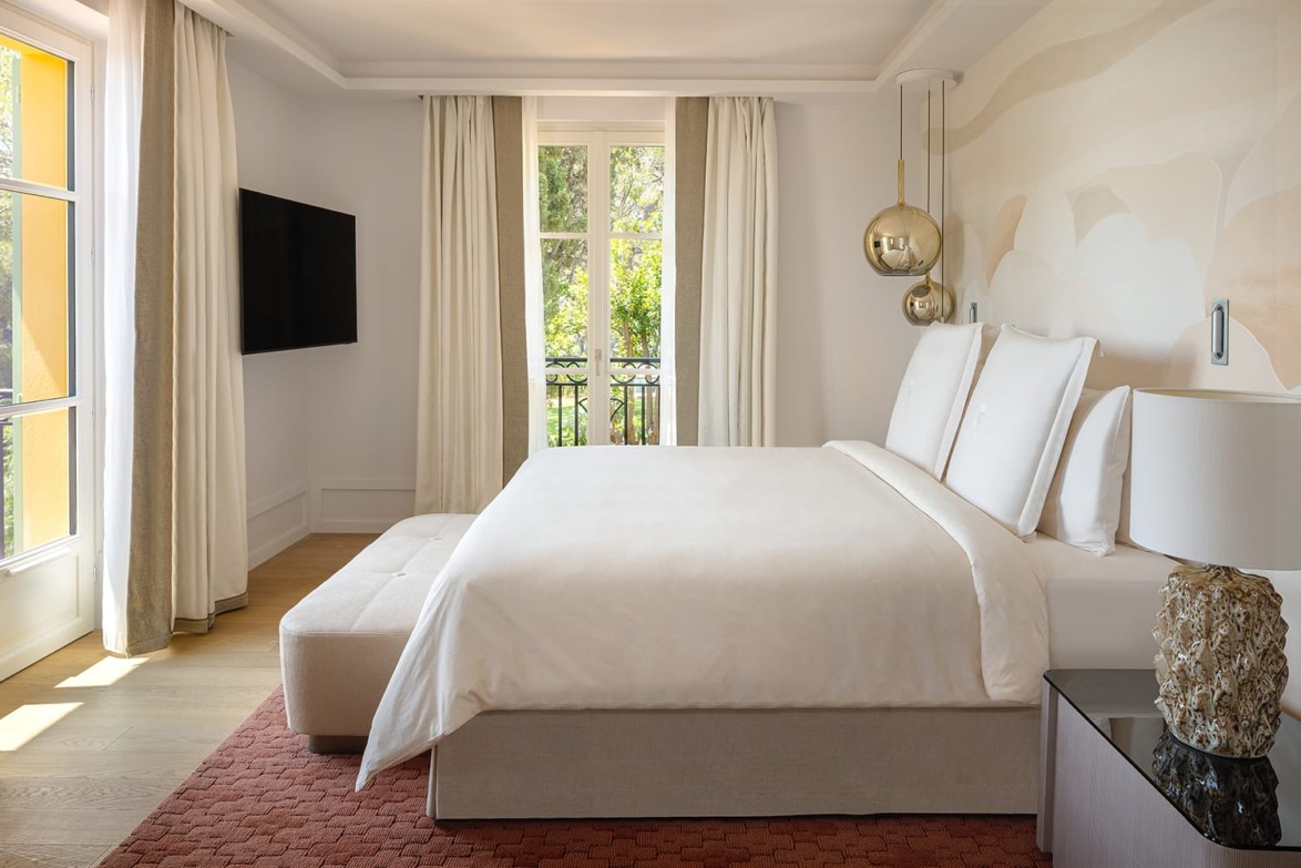 Modern hotel room with large bed, wall-mounted TV, and floor-to-ceiling windows with curtains, letting in natural light.