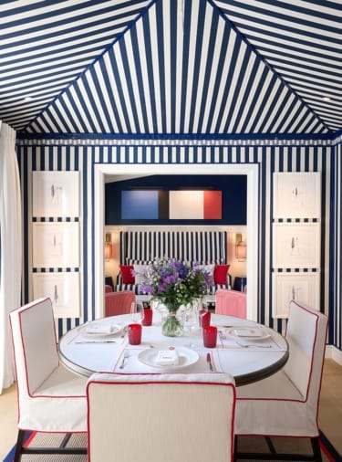 Dining room with blue and white striped walls and ceiling, round table set for four, pink chairs, and floral centerpiece.