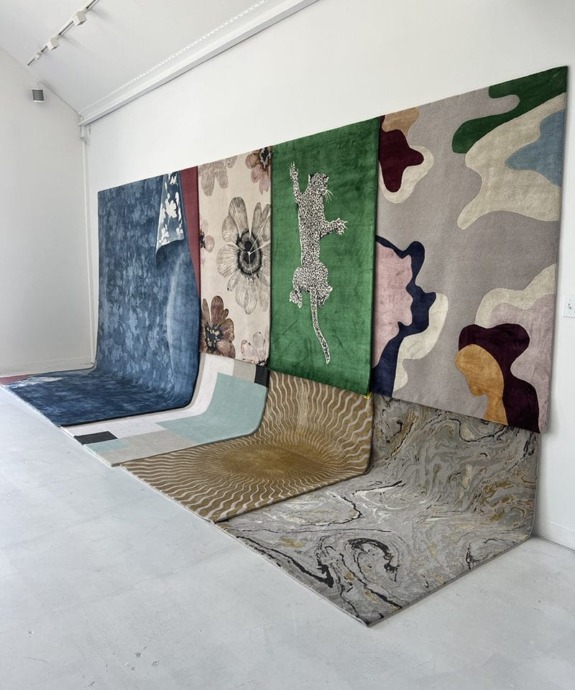 Assorted colorful rugs hung vertically in an art gallery with unique patterns, including florals and abstract designs.