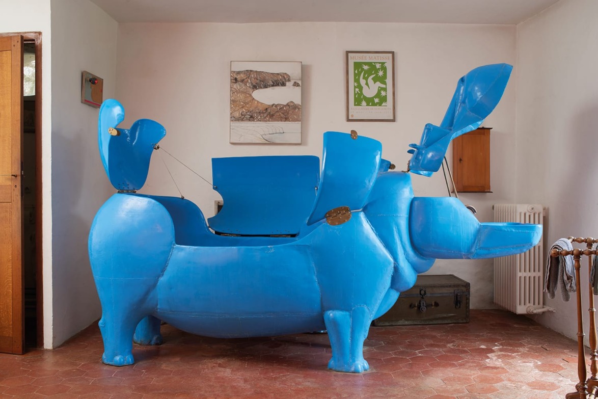 Large blue hippopotamus-shaped sculpture with open compartments in a room with artwork on white walls and terracotta floor.