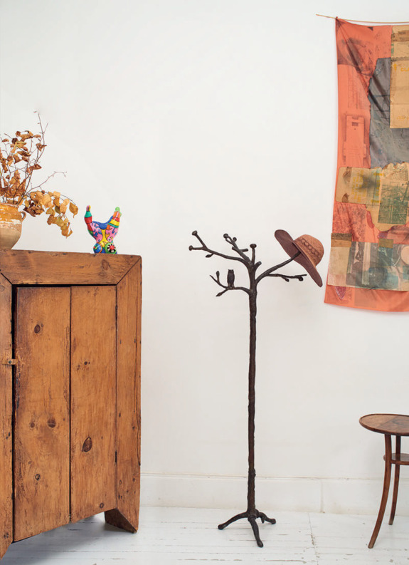 Vintage wooden coat rack with hat, beside a rustic cabinet and colorful wall art in a cozy room setting.