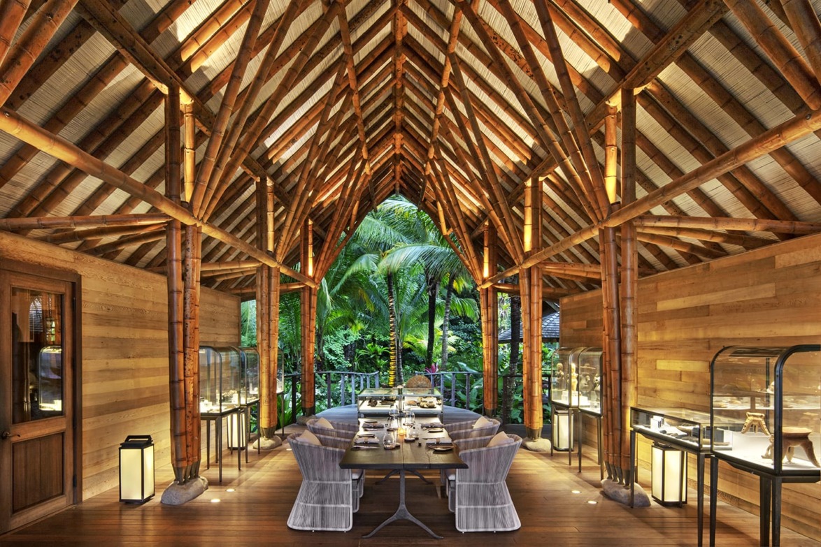 Elegant tropical dining area with wooden beams, glass display cases, and lush greenery visible through open archway.
