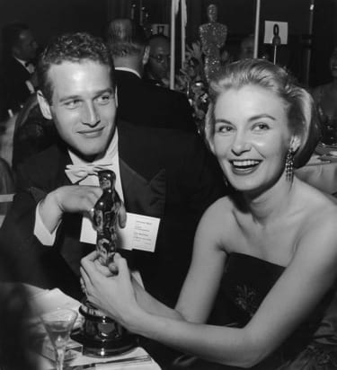 A smiling woman holding an award statue at a formal event, seated beside a man in a tuxedo.