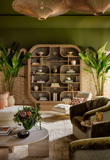 Cozy living room with green walls, large bookshelf, lush plants, and comfortable seating.