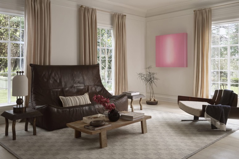 Modern living room with a leather sofa, wooden coffee table, large windows, beige curtains, and abstract pink artwork on the wall.