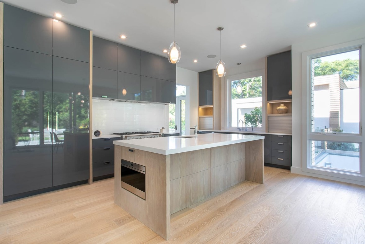 Modern kitchen with island, pendant lights, dark cabinets, large windows, and wooden flooring.