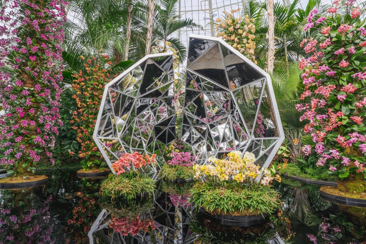Geometric sculpture surrounded by vibrant flowers and tropical plants in a greenhouse setting.