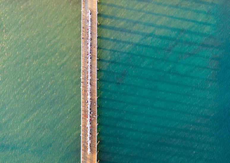 Aerial view of a pier extending over turquoise waters with people walking along it, creating long shadows in the sunlight.