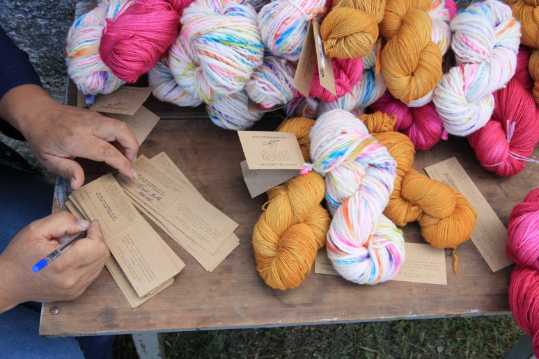 Person writing on tags next to colorful skeins of yarn on a table at an outdoor market.