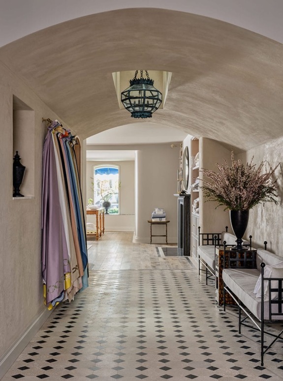 Elegant hallway with checkered floor, colorful hanging fabrics, a large vase with flowers, and a view into a sunlit room.