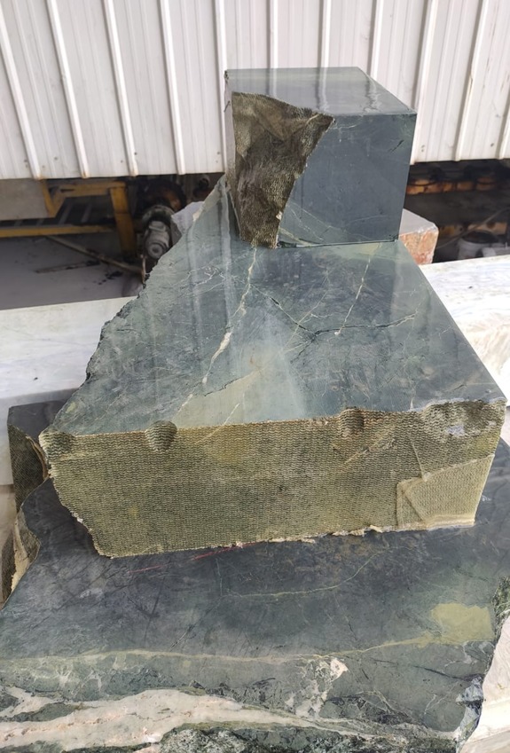 Block of natural stone and cube with polished and rough surfaces on a workbench near a white warehouse wall.