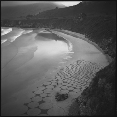 Black and white photo of a beach with geometric sand patterns and rolling hills in the background.