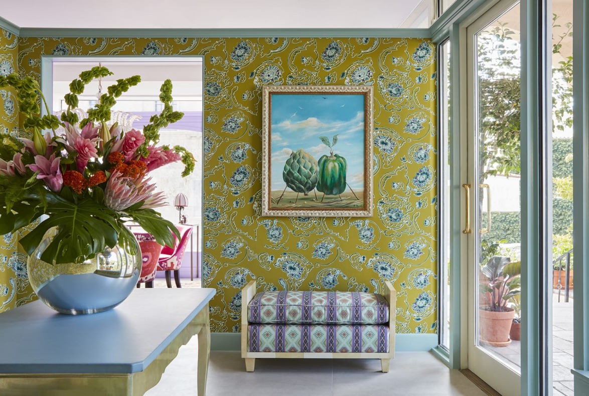 Floral wallpapered room with artwork, flowers in a vase, a patterned bench, and glass doors opening to a garden.