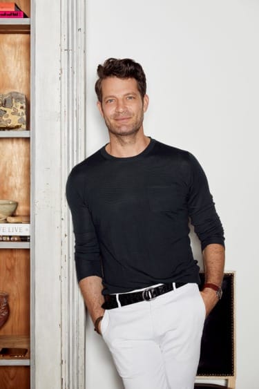 Person standing casually by a bookshelf, wearing a black long-sleeve shirt and white pants, with a relaxed expression.
