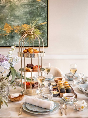 Afternoon tea setup with tiered tray of pastries, elegant tableware, flowers, and glasses of champagne on a decorated table.