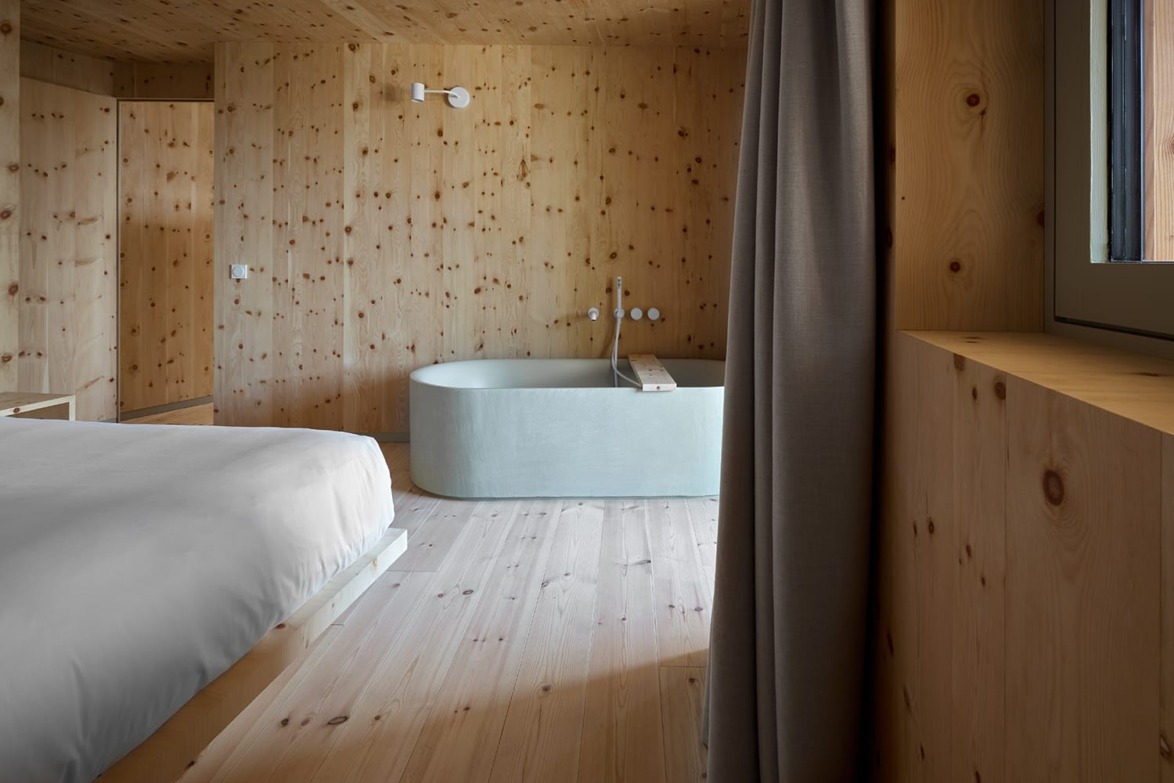 Rustic bedroom with wooden walls and floor, featuring a modern bathtub and a large window with gray curtains.