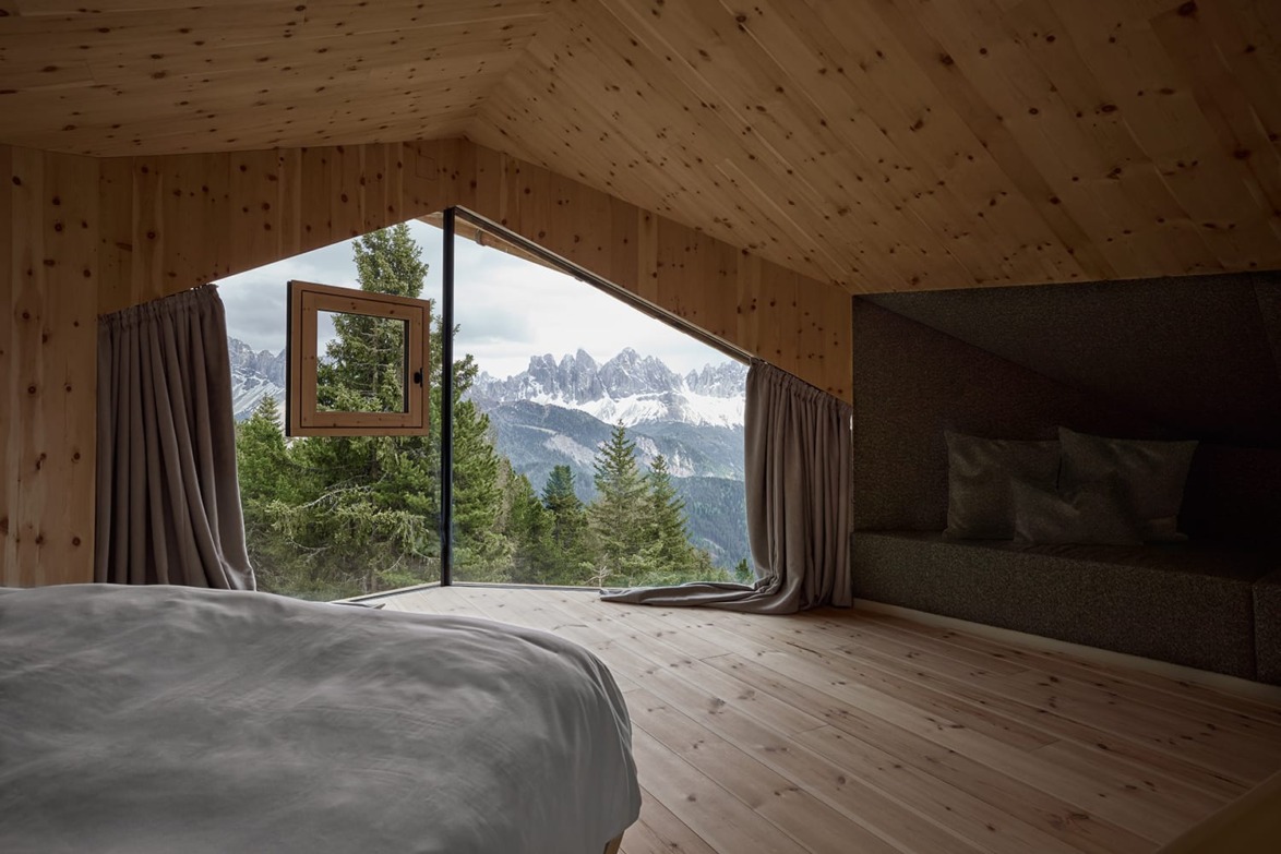 Cozy wooden cabin bedroom with large window view of snowy mountains and forest, showcasing serene natural landscape.