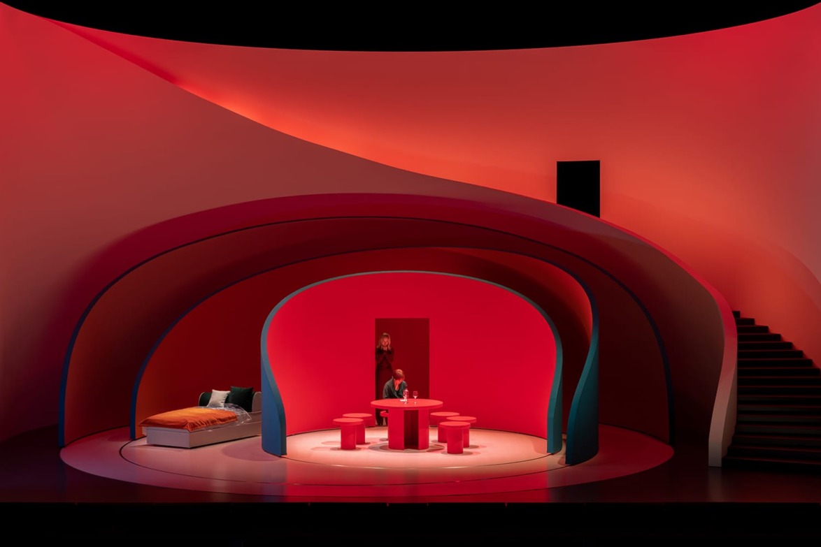 Modern stage set design with curved red walls, a central seating area, and stairs to the right, creating a dramatic ambiance.